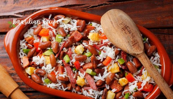 Clay baking dish of Brazilian rice and beans with diced peppers, corn, and ham, with a wooden spoon resting in it.