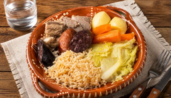 Clay plate with rice, pork chunks, chorizo, blood sausage, potatoes, carrots, and cabbage; glass of water nearby.