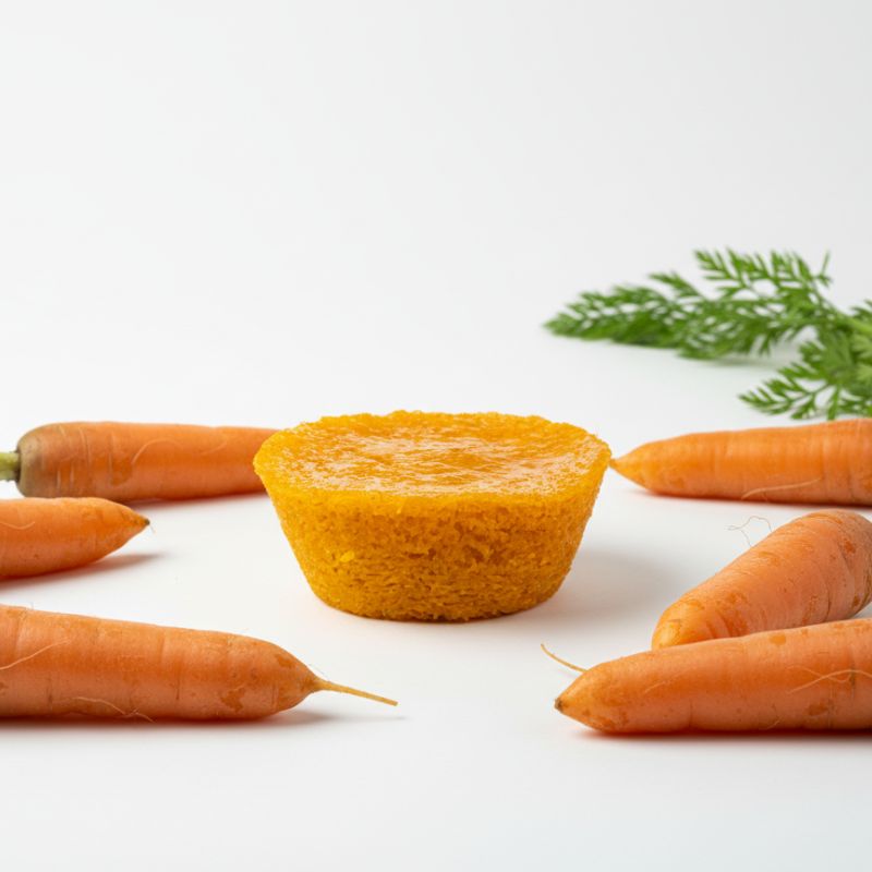 Queijada de Cenoura 2 - Delícias Orange carrot muffin at center, surrounded by whole carrots on a white background.