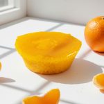Queijada de Laranja 2 - Delícias Bright yellow citrus tart on a white surface with a whole orange and orange segments nearby by a sunlit window.