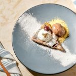 Dessert plate with a triangular pastry topped by vanilla ice cream and a cherry, dusted with powdered sugar on a blue plate.