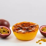 Queijada de Maracuja 2 - Delícias Yellow lemon tart with glossy passion fruit glaze on a white surface, flanked by halved passion fruits.