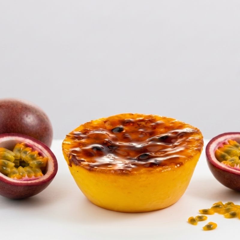 Queijada de Maracuja 2 - Delícias Yellow lemon tart with glossy passion fruit glaze on a white surface, flanked by halved passion fruits.