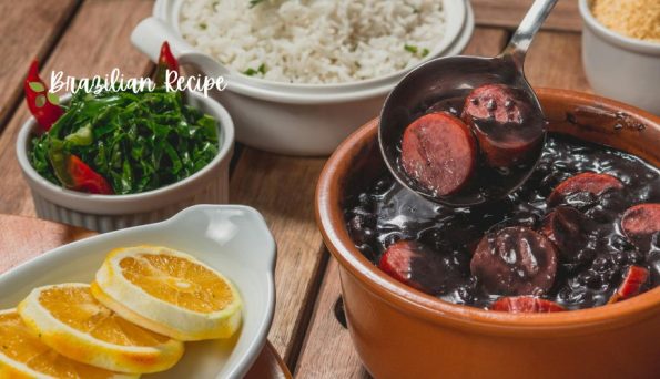 Feijoada (black bean stew) with sausage in a clay pot, plus orange slices, greens, and white rice on a wooden table.