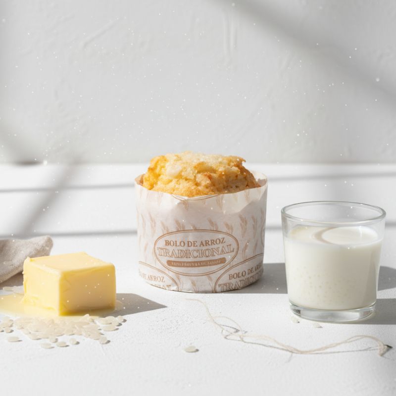 Bolo de Arroz - Delícias Traditional Brazilian rice cake in a paper wrapper, with butter cubes and a glass of milk on a white surface.