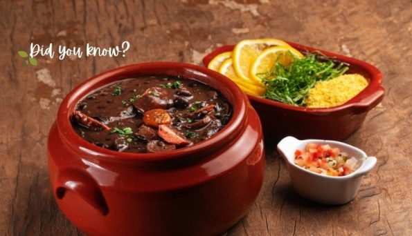 Red clay pot of black bean stew with sausage, garnished with herbs, next to lemon slices and salsa on a rustic wooden table.