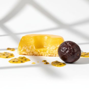 A slice of yellow cheesecake with a creamy center, plated with passion fruit sauce dots and a dark grape beside it on a white surface.