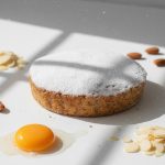 FRZB-PAT2947 - Delícias Round cake dusted with powdered sugar sits on a white surface, with a sunny-side raw egg yolk and almond slices nearby beneath bright light.