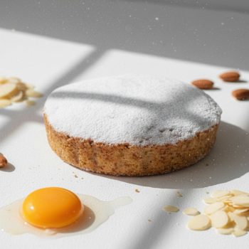 Round cake dusted with powdered sugar sits on a white surface, with a sunny-side raw egg yolk and almond slices nearby beneath bright light.