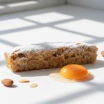 Tarte Santiago - Delícias Crumb cake slice dusted with powdered sugar sits on a white surface beside a bright orange egg yolk and a few almonds.