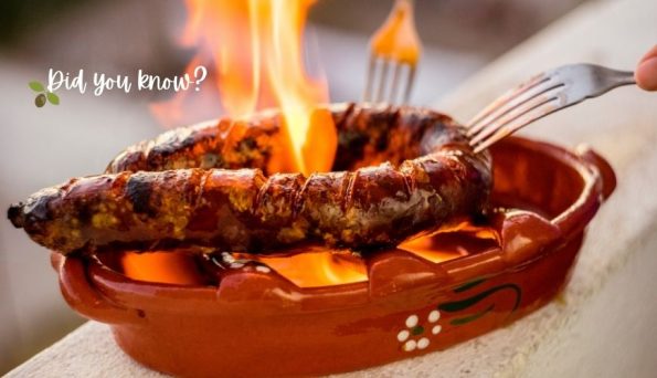 Sausage roasting on a clay grill over bright flames, being turned with a fork by someone off to the right.