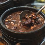 Brown beef bean stew in a dark clay pot, ladle lifting a chunky piece from the broth