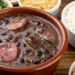 A clay pot of black bean soup with sausage slices and beans, with bowls of rice and toppings in the background.