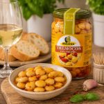 Jar of Maçarico Tremocos com Piri-Piri spicy chickpeas next to a bowl of chickpeas, with bread, a glass of white wine, garlic, and basil on a wooden board.