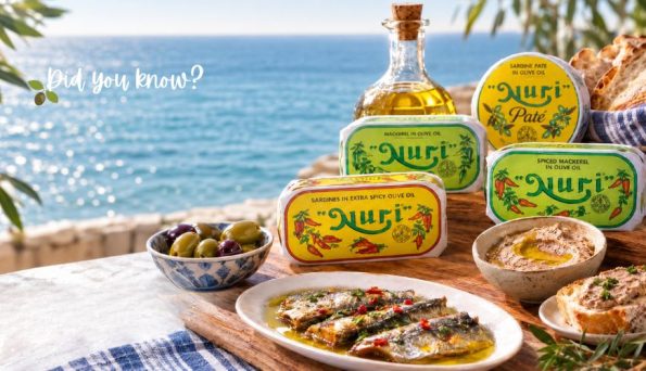 Seaside table with Nuri sardine tins, a bottle of olive oil, olives, bread, and a plate of sardines in olive oil by the coast.